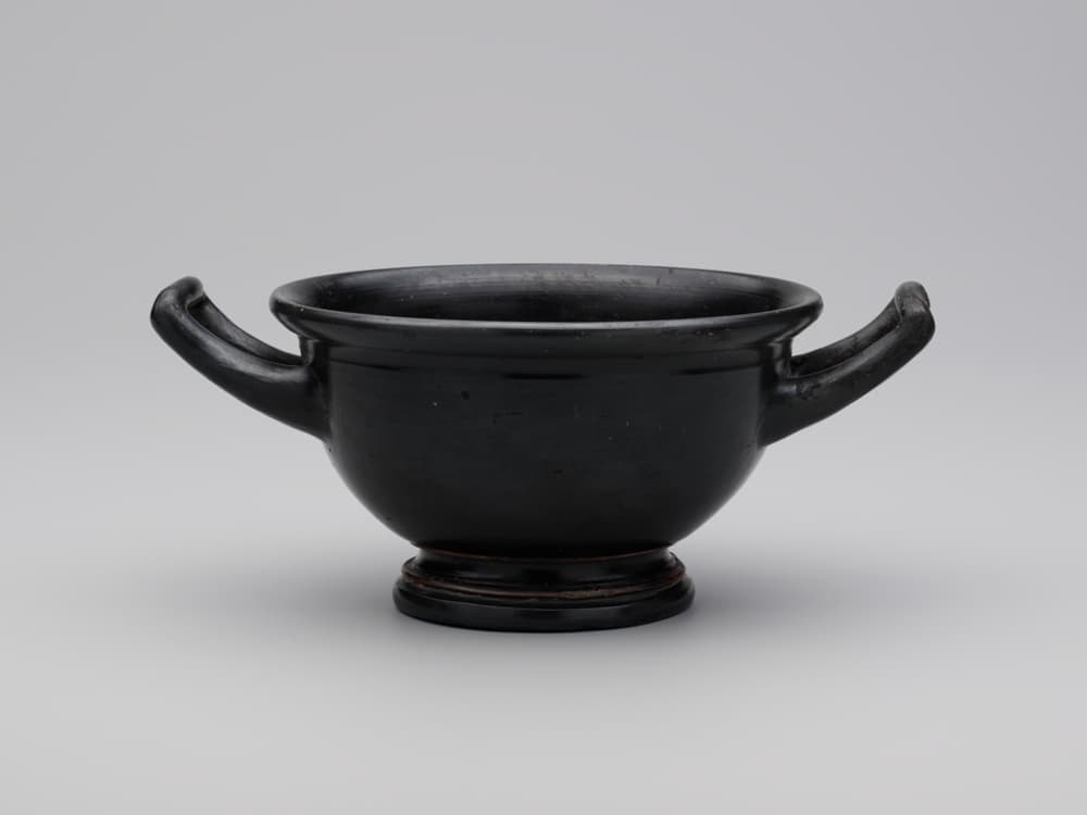 A wide-mouthed drinking cup glazed in black with two handles extending from opposite sides.