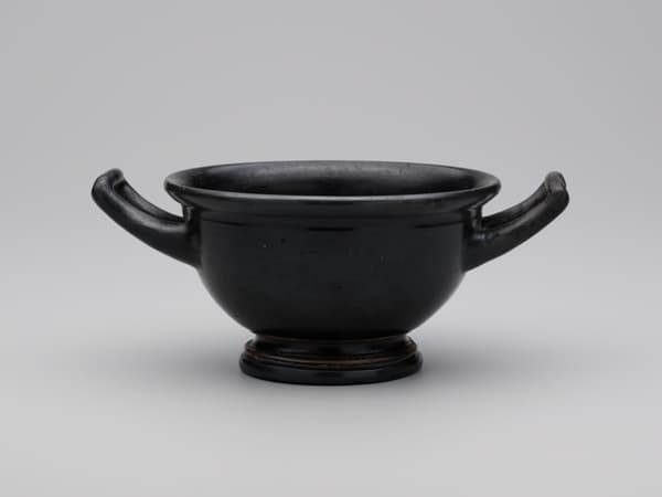 A wide-mouthed drinking cup glazed in black with two handles extending from opposite sides.